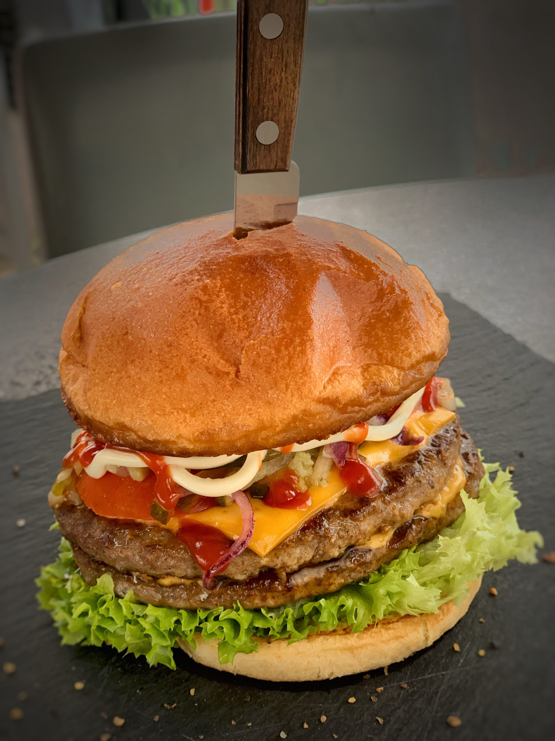 Hausgemachter Burger mit Käse und doppeltem Fleisch-Pattys. Ein Steakmesser steckt in der Mitte des Burgers, der auf einer Schieferplatte serviert wird.