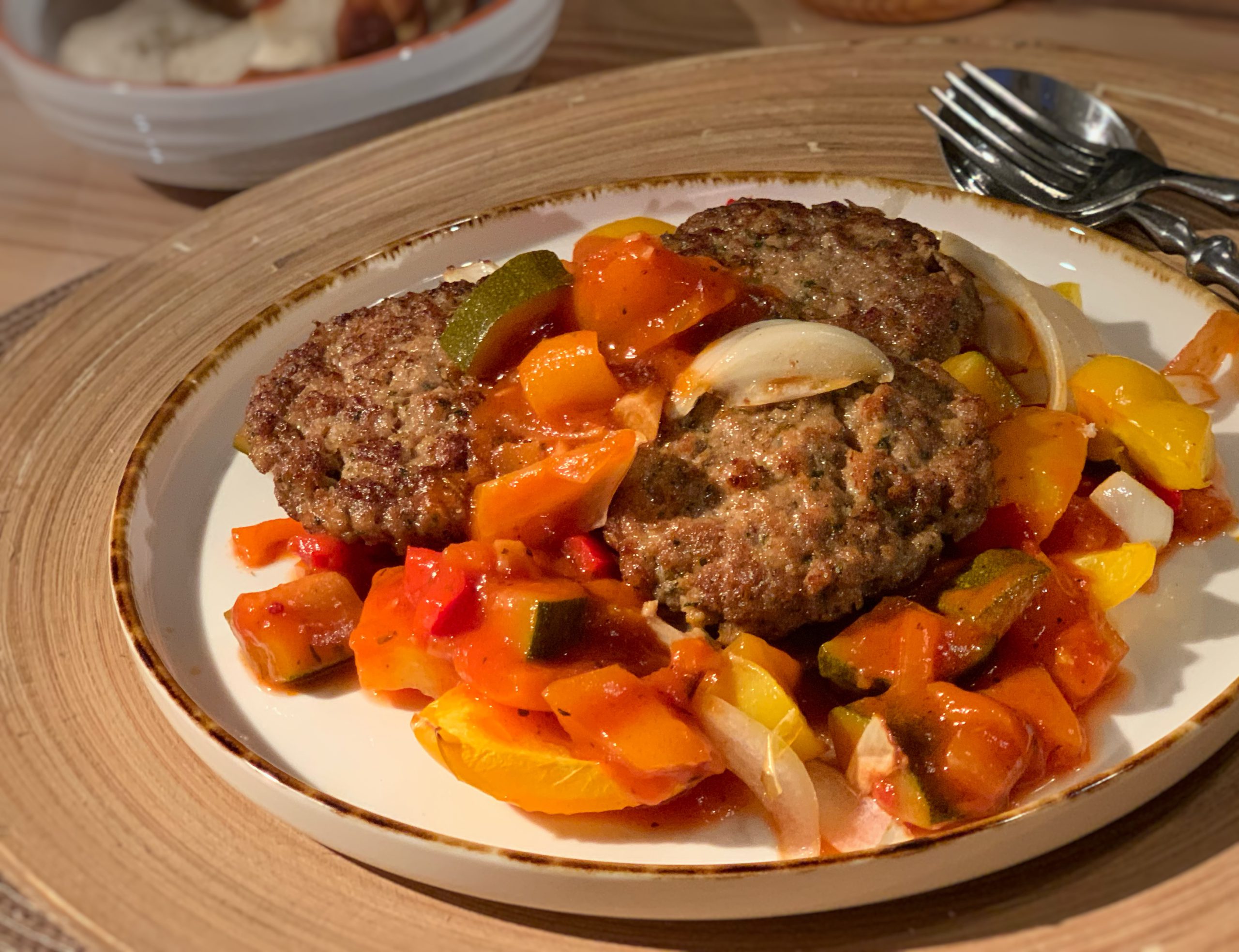 Fleischküchle mit Paprika-Zucchinisoße auf einem Teller. Daneben sieht man eine Gabel und einen Löffel.