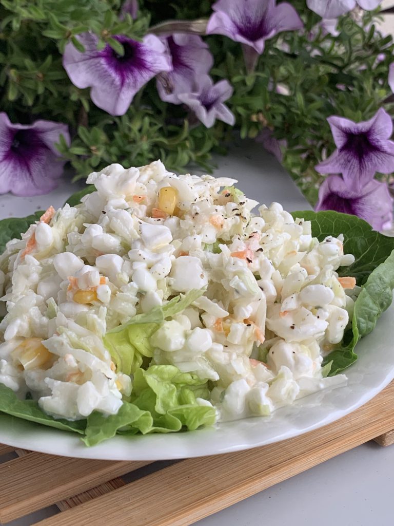 Krautsalat mit körnigem Hüttenkäse auf Salatblättern auf einem Teller. Hinter dem Teller sind lilafarbene Petunien zu sehen.