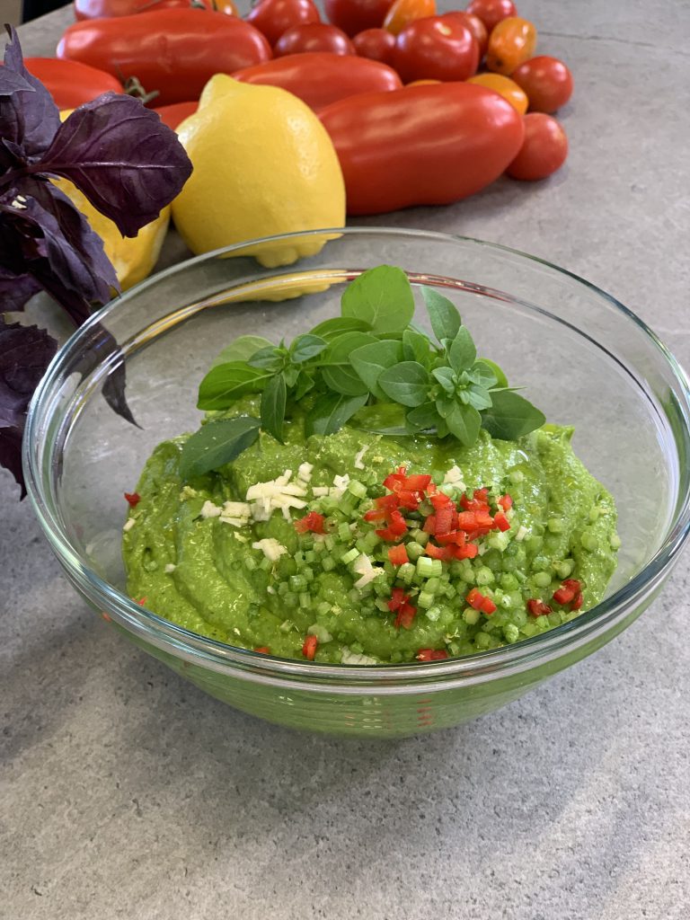 Basilikum-Zucchini-Dip in einer kleinen Glasschüssel. Der Dip wurde mit Basilikum-Blättchen und kleinen Würfeln von Paprika und Frühlingszwiebeln garniert. Die Schüssel steht auf einem grauen Untergrund und dahinter sieht man lilafarbene Basilikumblätter, zwei Ztronen und verschiedene Tomaten