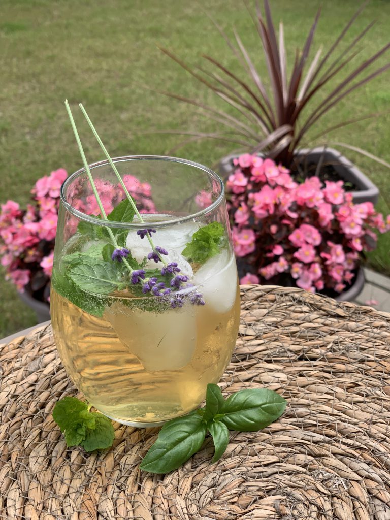 Birnen-Lavendelspritz in einem rundlichen Glas. Im Cocktail sind Eiswürfel, Minzblätter und zwei Lavendelblütenstängel drin. Das Glas steht auf einem Tisch, draußen im Grünen. Im Hintegrund sind rosa Blumen zu sehen.