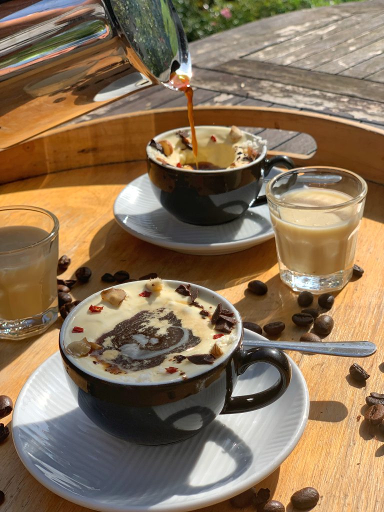 Auf einem Tablett sieht man zwei Tassen mit dem italienischen Dessert Affogato. Dekoriert sind die beiden Desserts mit Schokoladen- und Nusssplittern sowie Kakaopulver. Aus einer silbernen Kaffeekanne wird Espresso in die hintere Kaffeetasse gefüllt. Neben den Tassen stehen kleine Gläser mit Eierlikör. 