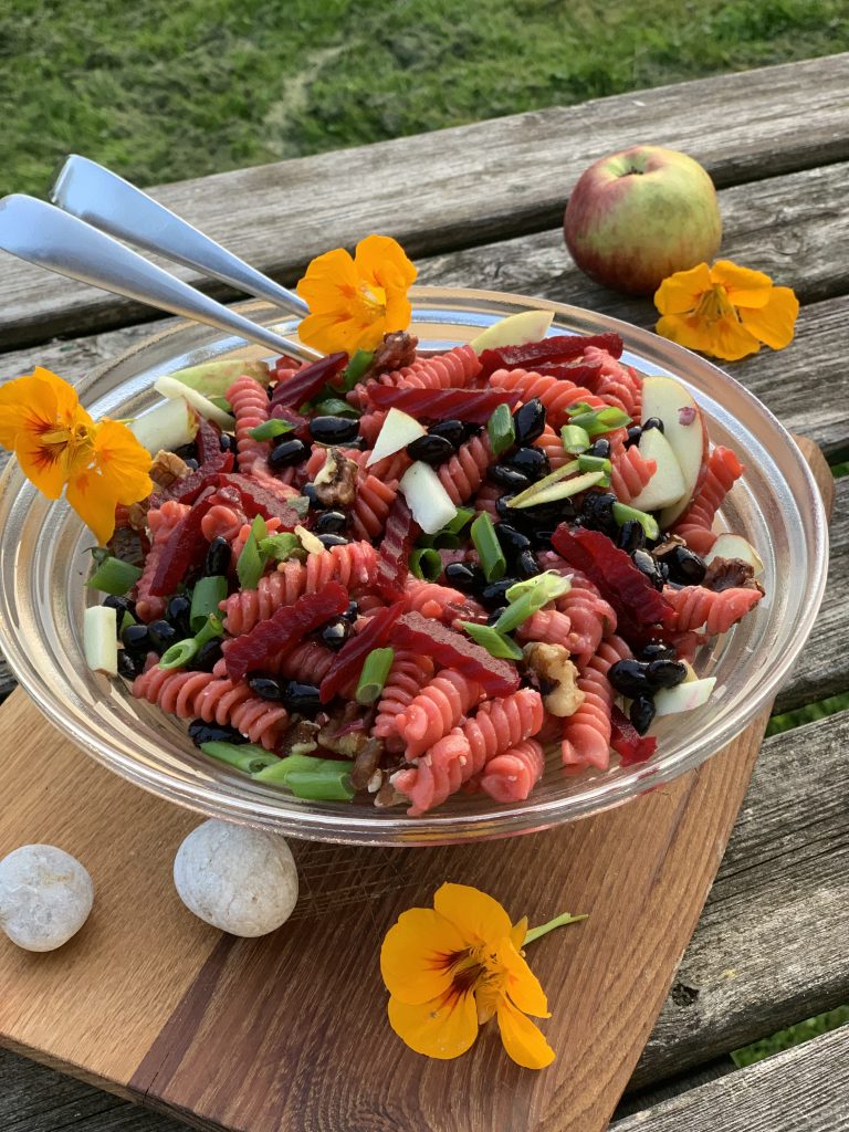 Man sieht eine große Glasschüssel mit Rote Bete - Nudelsalat mit Bohnen & Walnüssen. Die unterhalb der Schüssel befindet sich ein Holzbrett. Der Nudelsalat steht auf einem Holztisch, draußen im Gründen. Auf dem Tisch sieht man auch einen Apfel sowie vier Kapuzinerkresseblüten.