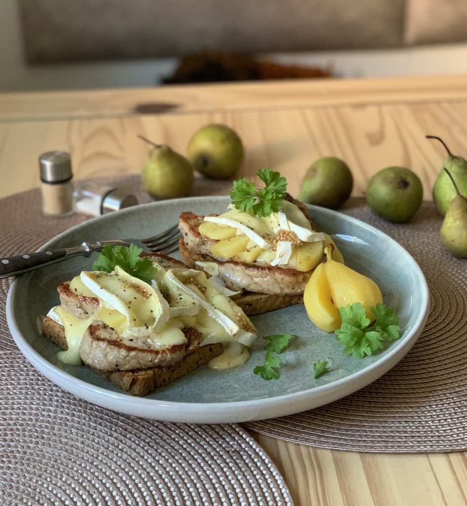 Zwei Schweinefilet-Toasts mit Currybirnen- und und Camembertscheiben auf einem grauen Teller. Der Teller steht auf einem Platzdekchen, der auf einem Holztisch steht. Auf dem Tisch sieht man auch einen Salz- und Pfefferstreuer sowie sechs Birnen. 