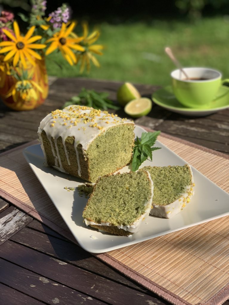 Auf einem quadratischen Servierteller liegt ein länglicher, angeschnittener Brennnessel-Limettenkuchen. Unter dem Servierteller sieht man ein Platzdeckchen aus Bambus. Neben dem Kuchen steht eine grüne Tasse mit Unterteller, zwei Limettenhälften und eine Vase mit verschiedenen gelben, lilafarbenen und rosanen Blumen. Das Arrangement steht draußen auf einem Holztisch. Im Hintergrund sieht man Gras.