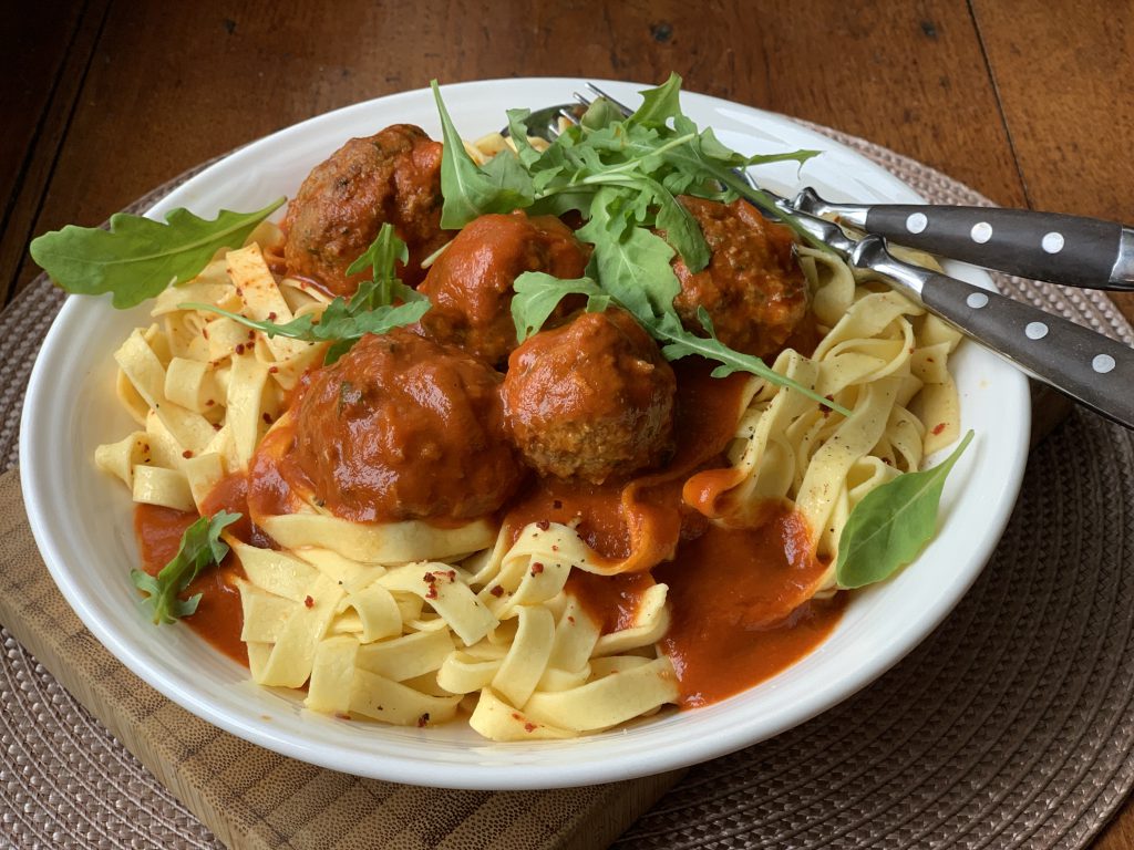 Ein Teller mit Bandnudeln mit Tomatensoße und Rindfleischbällchen. Über die Nudeln wurdenn Rucola-Blätter gestreut. Gabel und Löffel sind rechts am Telerrand sichtbar. Der Teller steht auf einem Holztisch.