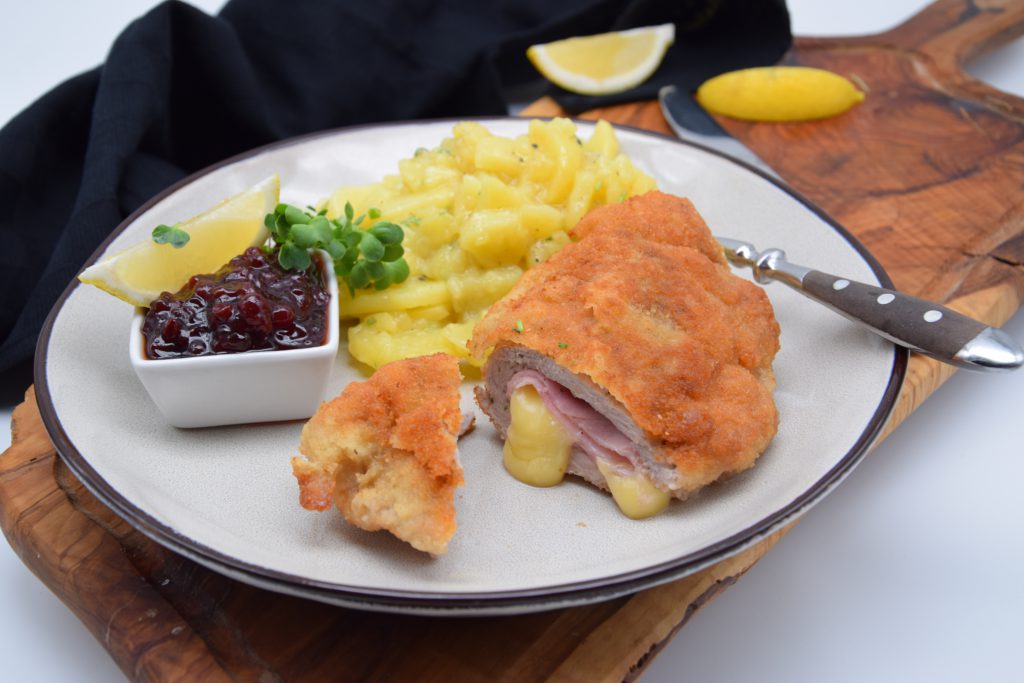 Ein knuspriges, goldenes Stück Cordon bleu, das aufgeschnitten ist. Der Käse fließt etwas aus dem größeren Teil heraus. Auf dem weißen Teller ist außerdem Kartoffelsalat, ein kleines Schüsselchen Preiselbeerkonfitüre, eine Zitronenspalte und als Dekoration etwas Kresse zu sehen. Eine Gabe liegt auf dem Tellerrand. Der Teller steht auf einem Holzbrett. Auf dem Holzbrett liegen auch 2 weitere Zitronenspalten und ein Messer. Im Hintgerund sieht man ein schwarzes Geschirrtuch.