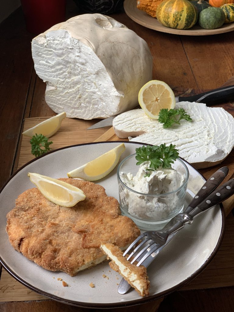 Ein angeschnittenes Schnitzel vom Riesenbovist auf einem Teller. Garniert mit zwei Zitronenspalten und Petersilie. Besteck und ein Gläschen mit Remoulade ist ebenfalls auf dem Teller. Neben dem Teller steht der angeschnittene Riesenbovist, eine halbe Zitrone, eine Zitronenspalte und im Hintergrund sieht man eine Schale mit Zierkürbissen.