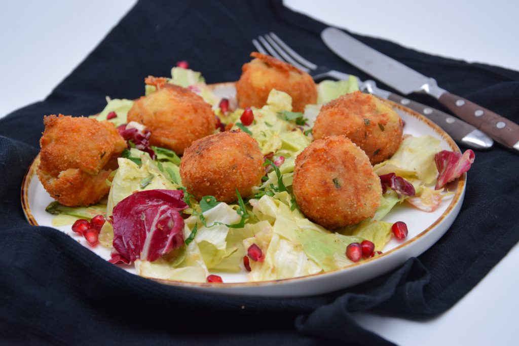 Sechs Kartoffelkäseknödel auf einem bunten Salatbett. Auf dem Salat sind Granatapfelkerne verteilt. Der Teller steht auf einem drappiertem, schwarzen Platzdeckchen. Neben der Teller sieht man Gabel und Messer.