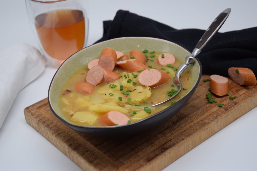 Saure Kartoffelrädle-Suppe mit, in Stücken geschnittenen, Saitenwürstchen. Die Suppe wurde mit Schnittlauchröllchen bestreut. Ein Löffel mit schwarzem Schaft liegt in der Schüssel, die auf einem Holzbrettchen steht. Hinter der Schüssel steht ein Glas mit Apfelsaft, eine schwarze Serviette und ein weißes Geschirrtuch. Auf dem Holzbrettchen sieht man auch zwei Stückchen der Saitenwürstchen und ein paar Schnittlauchröllchen.