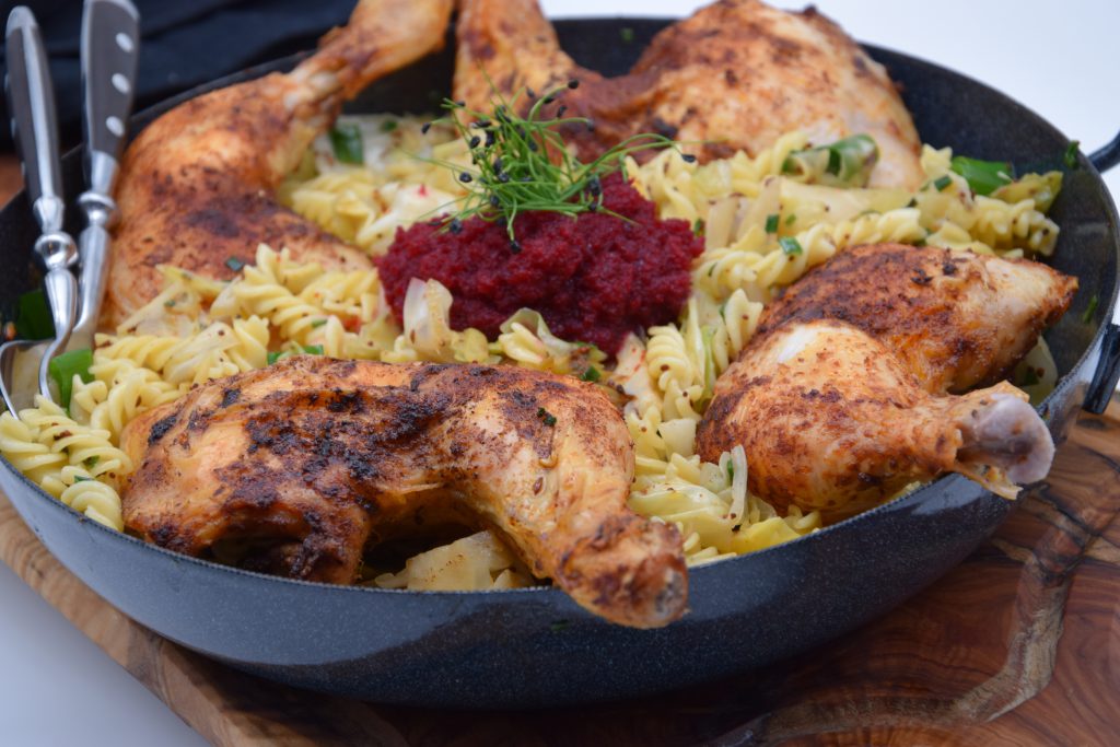 Vier Hähnchenkeulen in einer Pfnne mit Spitzkraut-Nudeln. Bei den Nudeln handelt es sich um Fusilli. Klein geschnittenen Kräuter wurden auf die Nudelpfanne gestreut. Die Pfanne steht auf einem Holzbrett, das auf einem weißen Untergrund steht. Ein Löffel und eine Gabel sind am Rand der Pfanne angelehnt. 