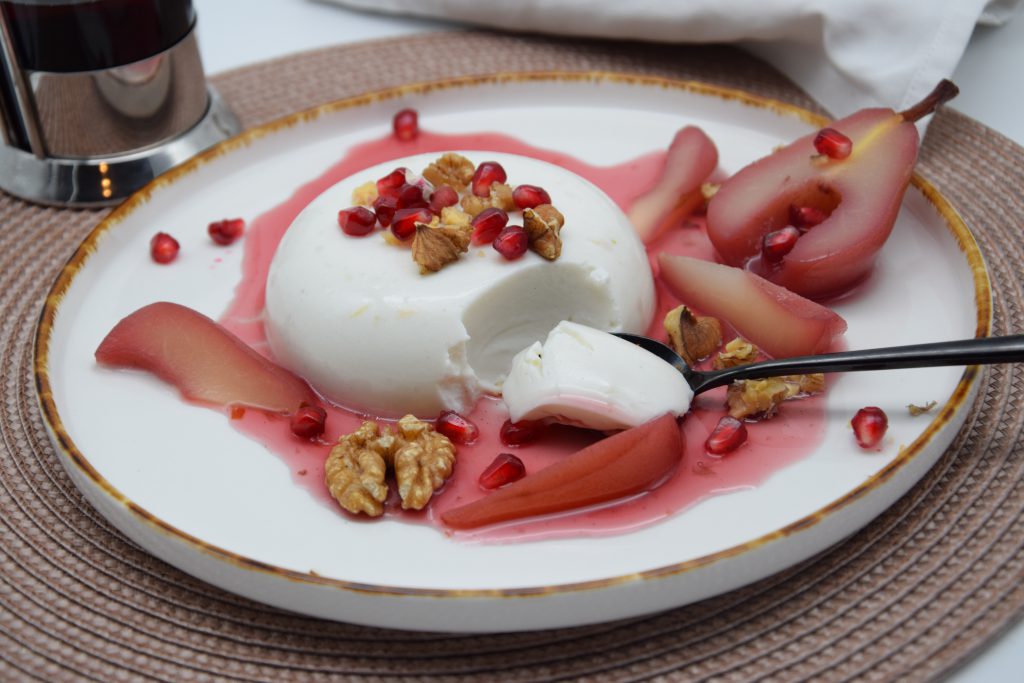 Ein runder Haselnusspudding in der Mitte eines flachen Tellers. Umrahmt mit Glühwein-Williamsbirnenhälften und Spalten. Das Dessert ist mit Granatapfelkernen und Walnussstücken garniert. Der Teller steht auf einem beigen Platzset auf einem weißen Tisch.