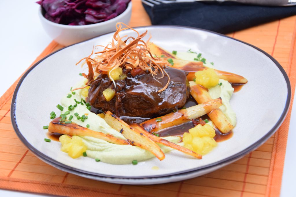 Hirschbraten mit klein geschnittenen Wurzelgemüsestückchen, Rosmarinbirnen und Bratensoße auf einem Teller. Dekoriert wurde das Gericht mit Schnittlauchröllchen und Karottenspänen. Der Teller steht auf einer Bastmatte. Dahinter kann man teilweise ein kleines Schüsselchen mit Rotkohl erkennen.