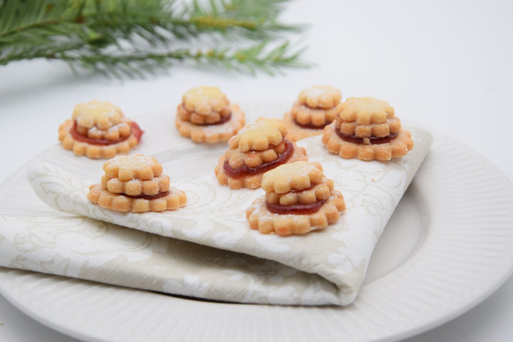 Sieben Terassenplätzchen auf einer weißen Stoffserviette, die auf einem Teller platziert ist. Im Hintergrund sieht man einen Tannenzweig. Der Teller steht auf einem weißen Tisch.