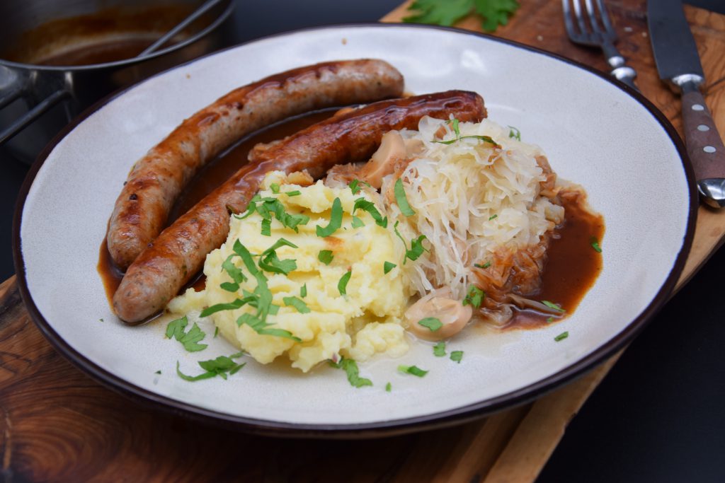 Zwei Bauernbratwürste mit Kartoffelpüree, Sauerkraut und braune Sauce auf einem hellen Teller. Das Püree ist mit geschnittener Petersilie bestreut. Neben dem Teller sind Messer und Gabel. Der Teller steht auf einem brauene Holzbrett.