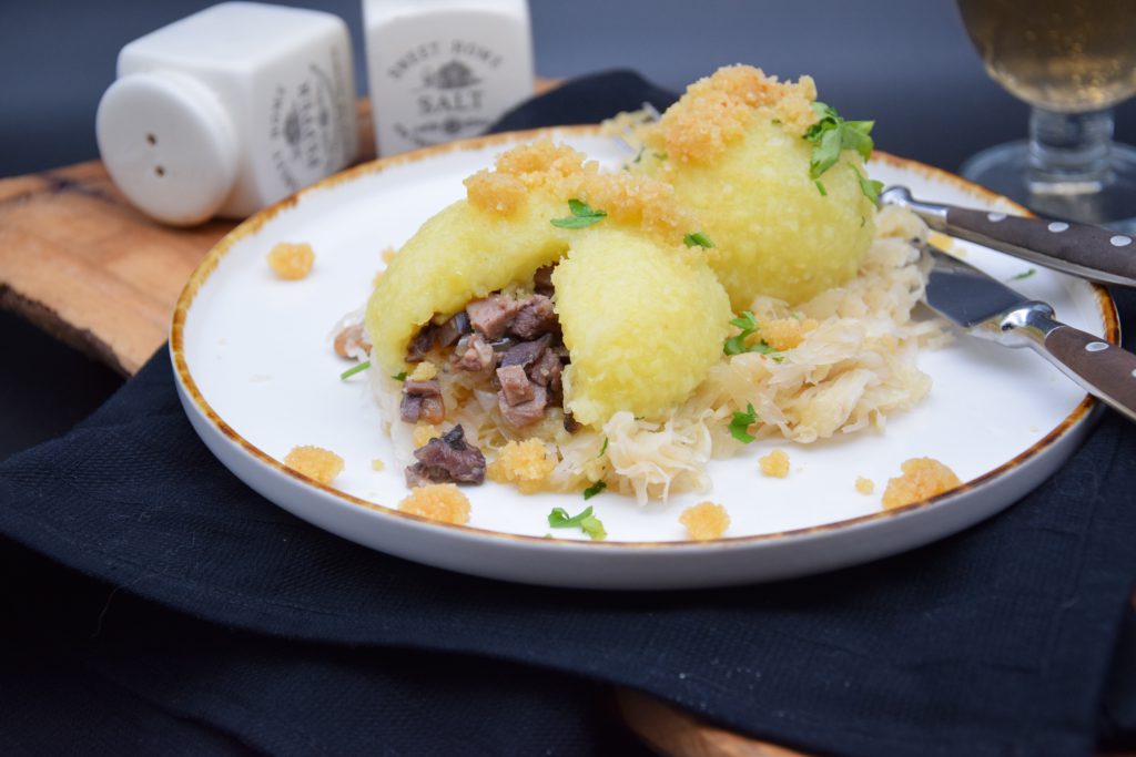 Auf einem hellgrauen Teller sieht man zwei gefüllte Kartoffelknödel auf Sauerkraut. Ein Knödel ist ausgeschnitten und etwas Füllung ist zu sehen. Auf dem Teller sieht man auch Kleckse von Semmelbröseln. Der Teller steht auf einer schwarzen Stoffserviette. Messer und Gabel sind auf dem Tellerand zu sehen. Im Hintergrund sieht man am oberen linken Bildrand einen Salz und Pfefferstreuer. 