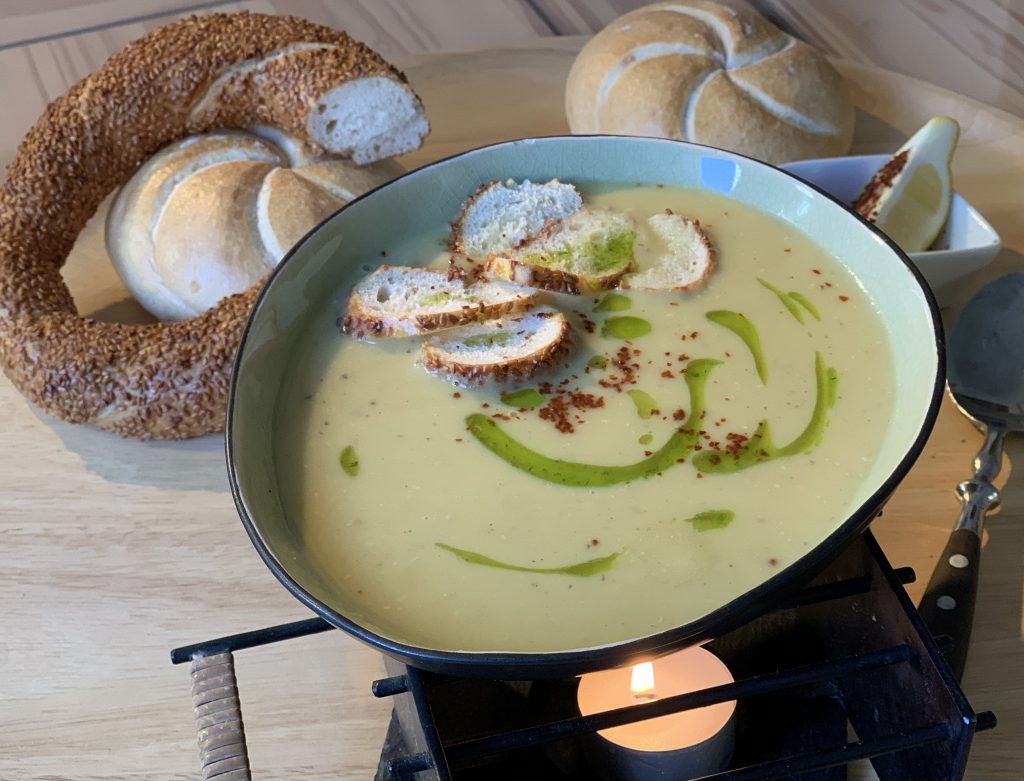 Linsensuppe in einem Suppenteller. Die Suppe wurde mit ein paar Scheiben von einem Sesamkringel sowie etwas Paprikapulver und Minzöl garniert. Neben dem Teller liegen der angeschnittene Sesamkringel, zwei Brötchen, ein kleines viereckiges Gefäß mit einem Zitronenviertel und einem Löffel. Der Teller steht auf einem Stövchen. Man sieht auch das Teelicht. Alles steht auf einem Holztablett.