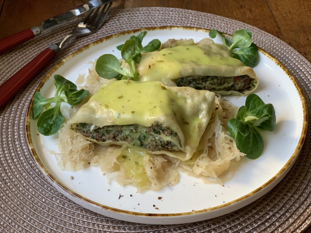 Ein weißer Teller mit zwei Maultaschen auf Sauerkraut. Dekoriert mit 5 Feldsalatbüscheln. Der Teller steht auf einem beigen Platzdeckchen. Links neben dem Teller liegen Messer und Gabel.