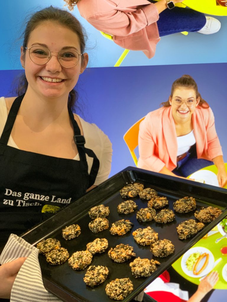 Ramona Kreuzer präsentiert ein Backblech mit Keksen. Sie hat eine schwarze Kochschürze an, auf der, der Slogan " Das ganze Land zu Tisch" steht. Sie trägt eine Brille und hat das Haar zu einem Zopf zusammengebunden. Im Hintergrund sieht man ein Plakat mit blauemm Hintergrund, auf dem sie an einem Tisch sitzt. 