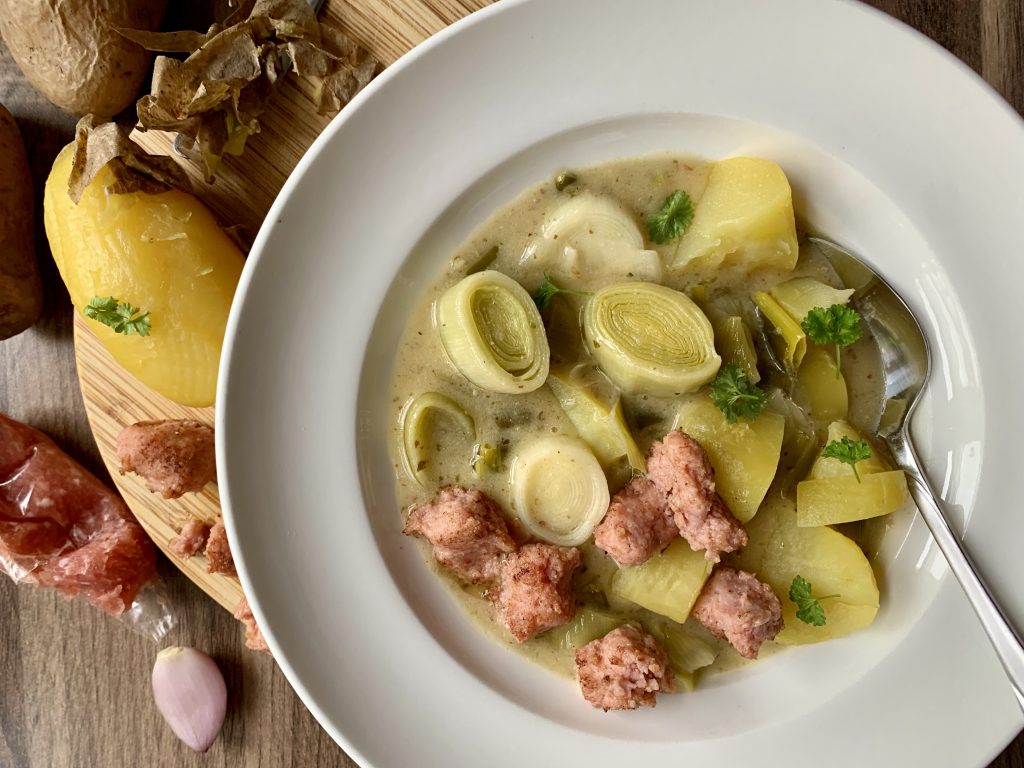 Rahm-Lauch mit Kartoffeln und Zwiebelmettwurststückchen in einem Suppenteller. Garniert ist das Essen mit geschnittener Petersilie. Ein Löffel liegt in dem Eintopf. Neben dem Teller sieht man eine ganze Kartoffel und die Mettwurst sowie eine Knoblauchzehe. Der Teller steht auch einem Holzbrettchen.