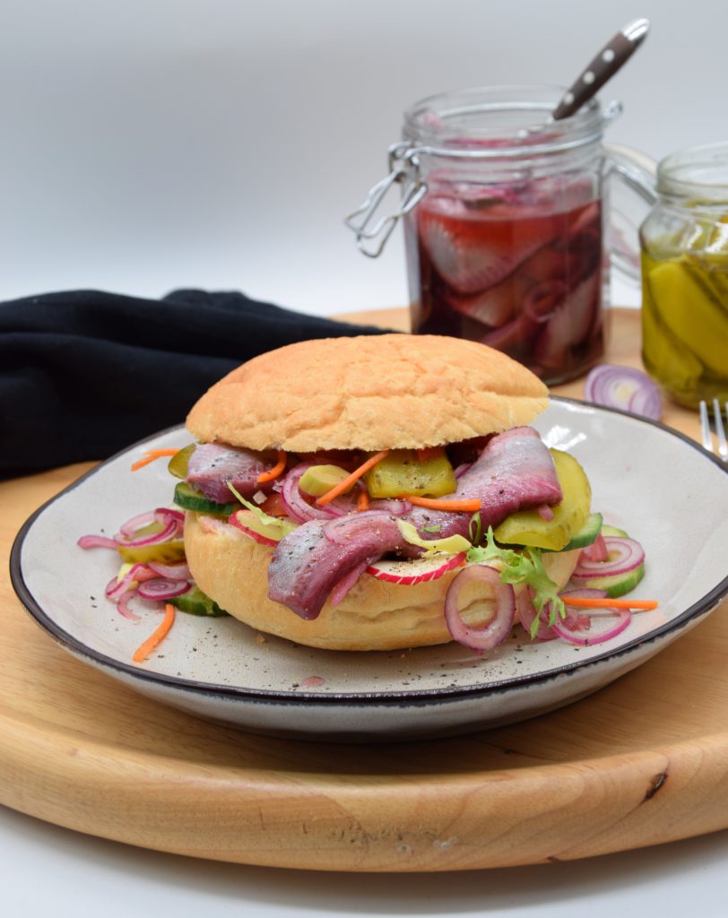 Ein helles Brötchen geüllt mit Rotweinmatjesfilets, roten Zwiebelringen, Karottenstiften, Salat, Radischenscheiben und eingelegte Gurkenscheiben. Das Brötchen ist auf einem weißen Teller mit schwarzem Rand, der auf einem hellbraunem Holztisch steht. Hinter dem Teller stehen zwei offene Einmachgläser. Eines ist gefüllt mit dem Rotweinmatjes und das andere mit Gurkenscheiben. Im Glas mit dem Matjes steckt auch eine Gabel. Links am Bildrand sieht man ein schwarzes Geschirrtuch.